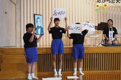 舞台の前で肖像画をもって立っている生徒、「シューベルト」と書かれた紙を頭上に掲げている生徒、「シューマン」「ブラームス」と書かれた紙を持っている生徒、舞台上で正座をして座っている生徒の写真