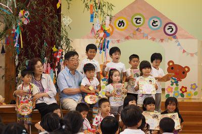 お誕生日の子供たちが手作りのメダルや本を持って前に並び高齢者の方と記念写真を撮っている写真