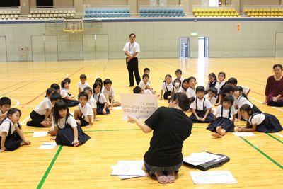 子供達が床に座り先生が紙で説明している様子を子供達が聞いている写真