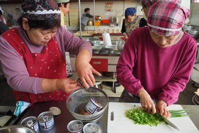 1人の女性が缶を開けザルの上で水切りをし、1人の女性がその横でねぎを刻んでいる写真