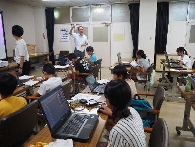 講師の方が前に立ち、右手をあげ子供達に話しかけていて、子供達は机に置かれているパソコンを見ている写真