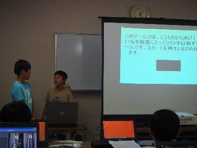 スクリーンでゲームに関しての説明文が映し出され、その隣で男の子2人が発表している写真