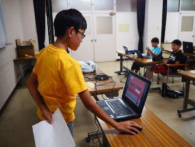 黄色いシャツに眼鏡をかけた男の子が教室の前に立ち、マウスを触りながら画面を見ている写真