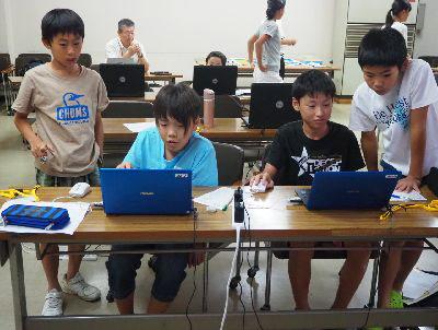 青いパソコンを2人の男の子が使っています。その作業している姿を横で立ちながら見ている男の子たちの写真