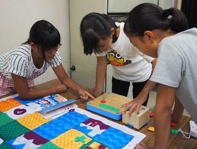 机で女の子3人が緑・赤・青の小さい飾りをボードの上に考えながら置いている写真