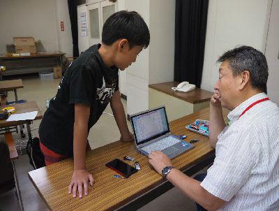 先生がパソコンを使って説明しています。男の子がパソコンの画面を見ながら先生のお話を聞いています。