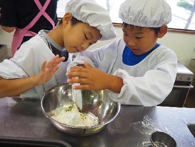 給食着を着た男の子2人が、ボールに入った粉の中に粉茶を入れている写真