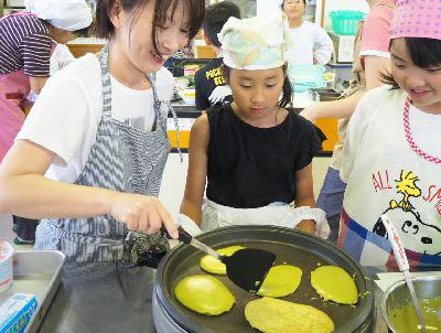 ホットプレートで焼いているパンケーキを、フライ返しひっくり返そうとしている女性と、その横で見ている女の子2名の写真
