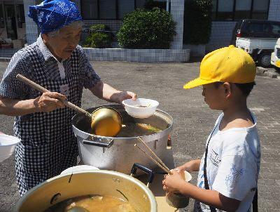 年配の女性が男子児童に鍋から豚汁をお玉ですくってお皿についであげている写真