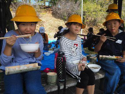 女子児童が竹の器に入ったご飯をひざに乗せながら、豚汁を食べている写真
