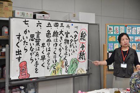 講師の藤田 正子さんがホワイトボードに貼られた絵手紙を描く7つのポイントが書かれた紙を指し棒で指しながら話をしている写真