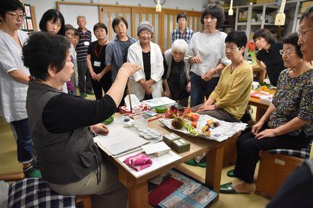 右手で筆を持ち机に座っている講師の藤田 正子さんの周りに受講生たちが集まり見ている写真