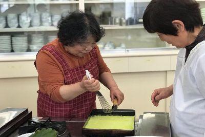 女性が卵焼き器で焼いた緑色の生地をフライ返しで取ろうとしている写真