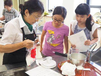 角田 眞理子さんが参加者の女の子2人と、調理台の上で砂糖の分量を量っている写真