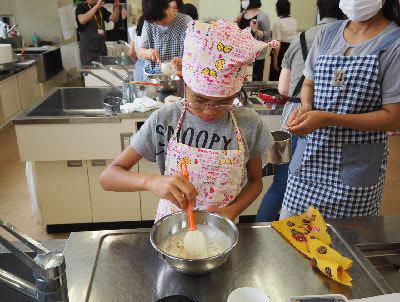 調理台の上でエプロンをつけて三角巾を頭に巻いた女の子がボールを持ってゴムベラでケークサレの生地を混ぜている写真