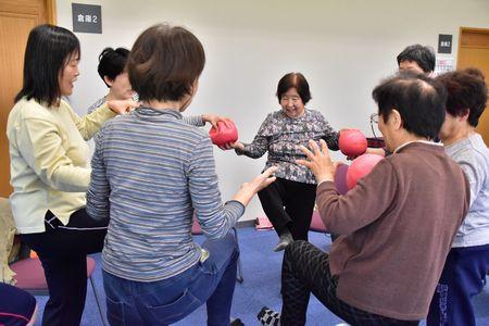 参加者の人で円になり、片足を上げながらピンクの柔らかいボールを隣の人に渡そうとしている写真