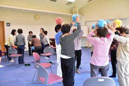 グループで円になり立ち上がりながら水色や黄色赤の柔らかいボールを頭に乗せている写真