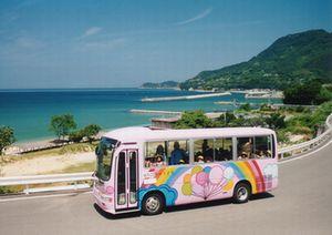 海岸沿いの道路を、側面にピンク色を基調としたイラストが描かれているバスが走っている写真